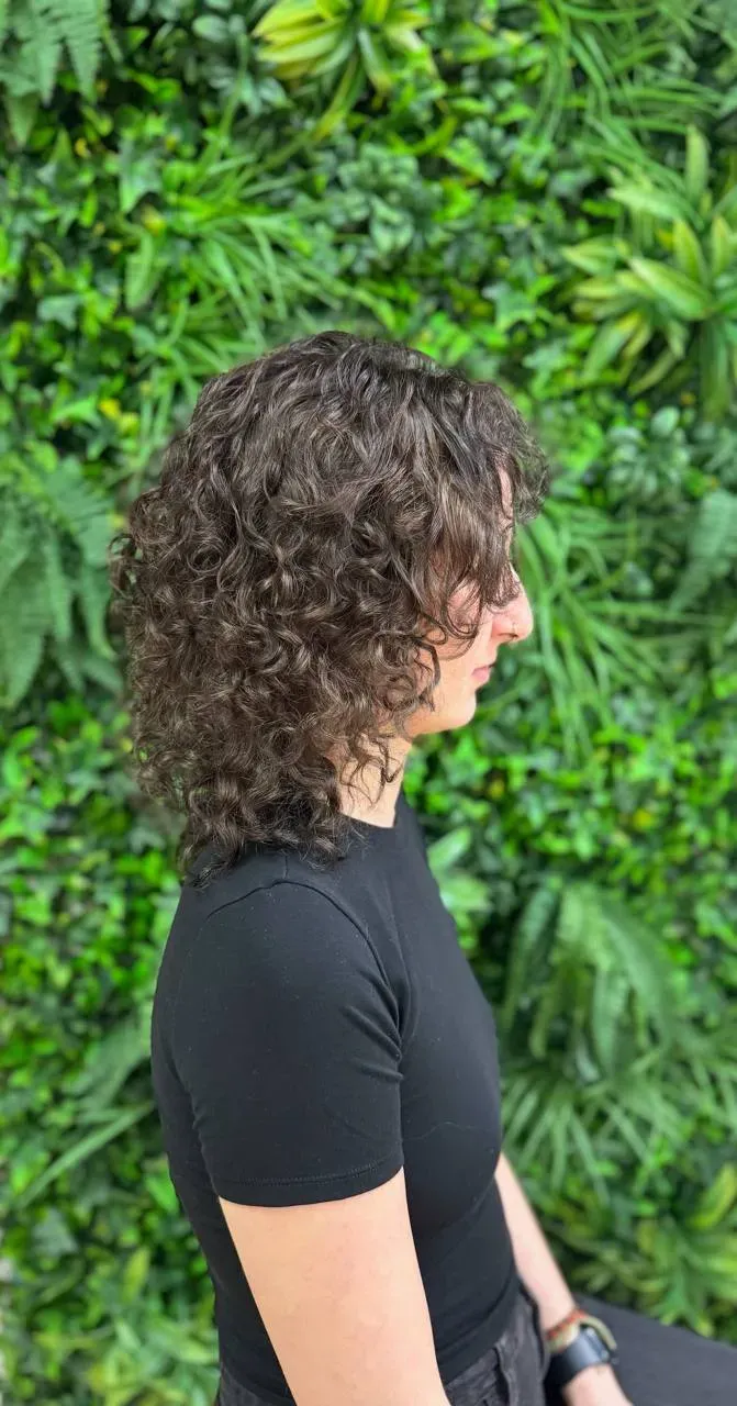 Frau mit voluminösen, dunklen Locken im Profil vor grünem Hintergrund – Ergebnis einer Dauerwelle bei Chic & Chill.